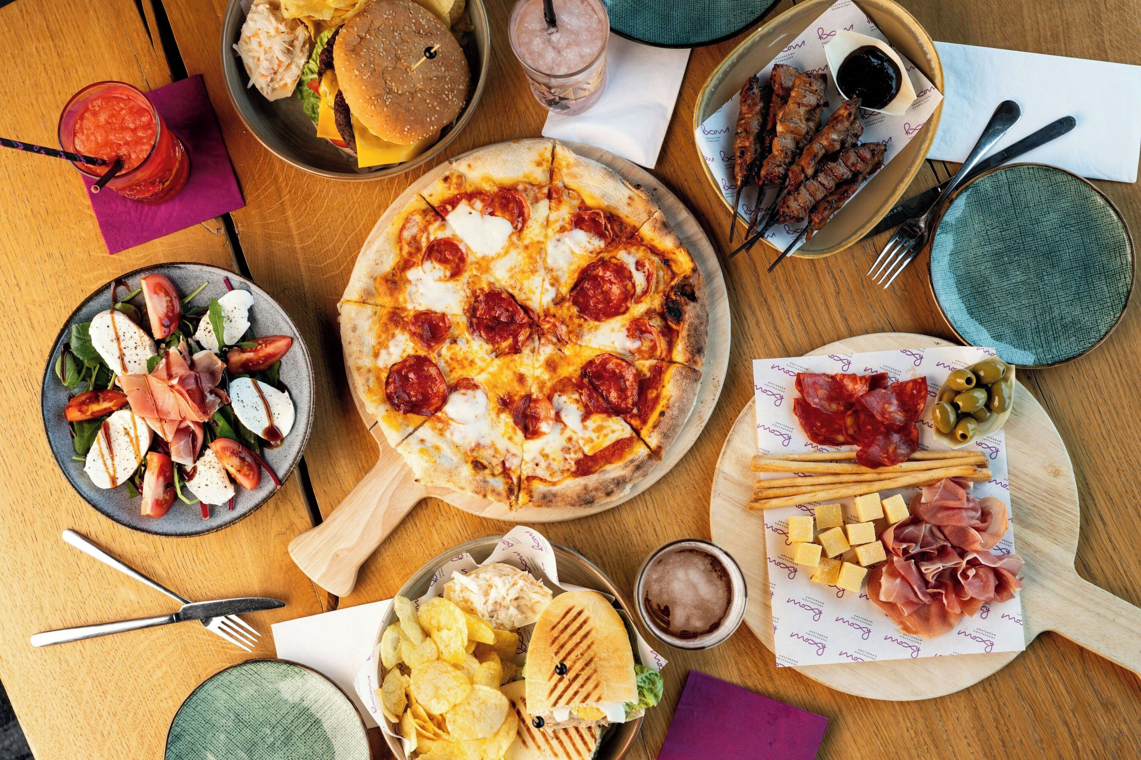 A variety of food items including pizza, salad, grilled meat, and chips on a wooden table.
