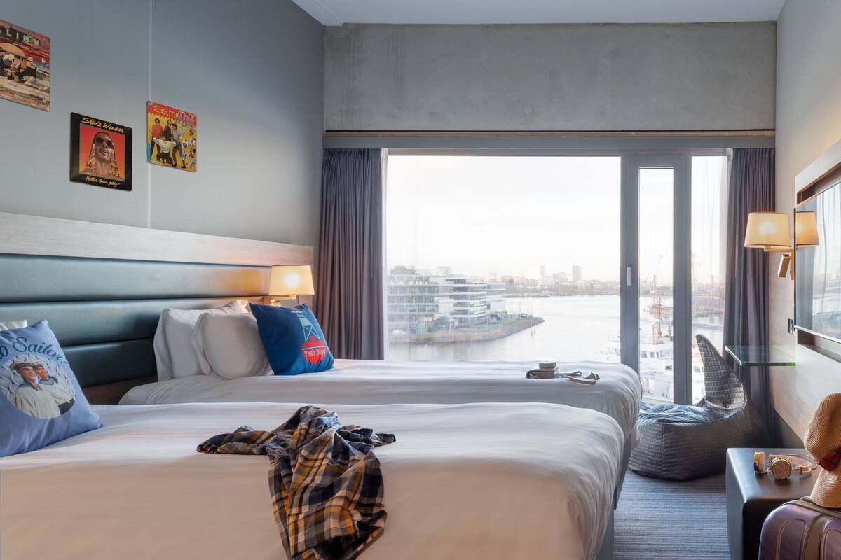 Quarto de hotel com duas camas, vista para o horizonte urbano e uma grande janela com cortinas.