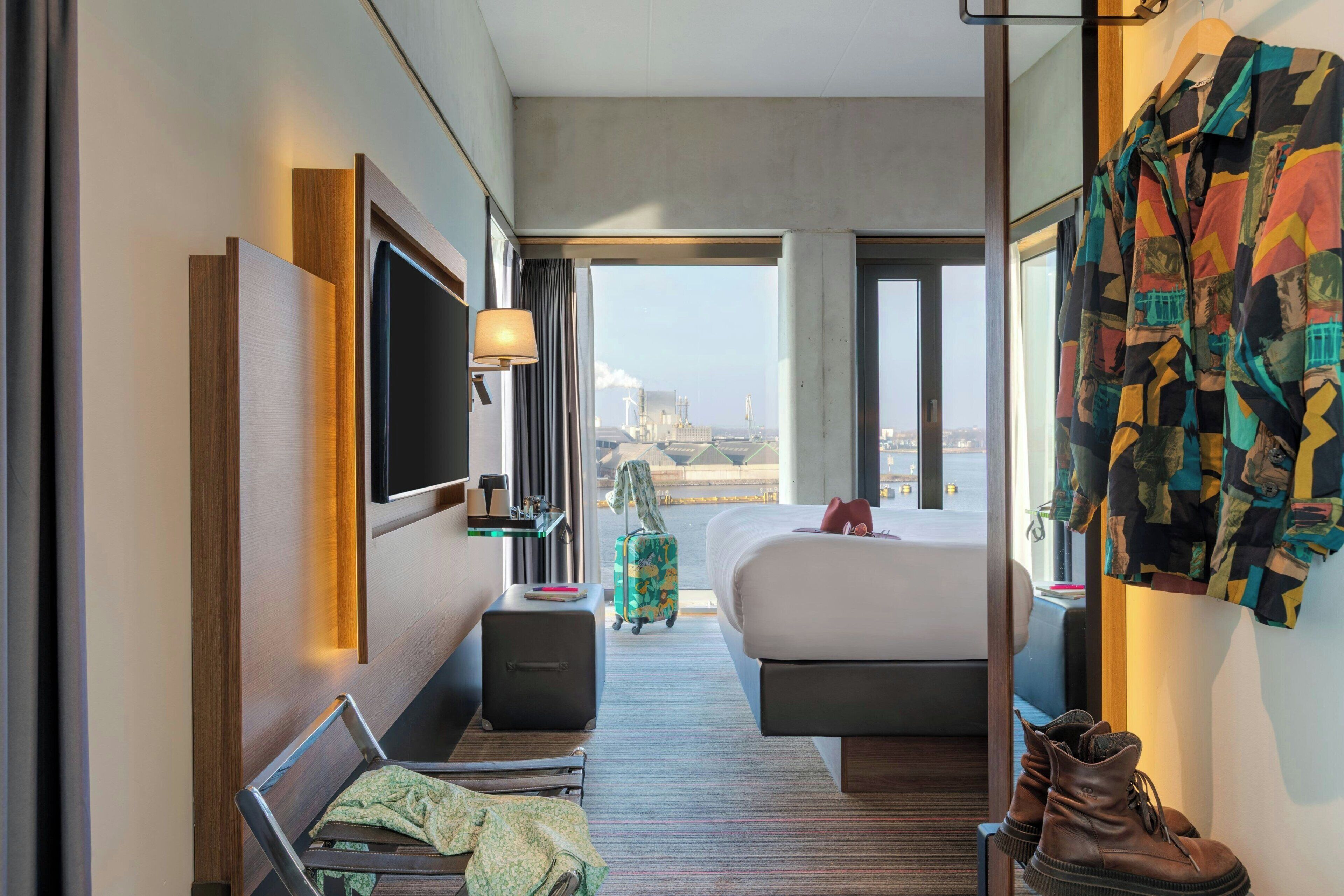 Une chambre d’hôtel comprenant un lit, un bureau, une chaise, une télévision, une fenêtre donnant sur la ville et un porte-manteau avec une veste colorée.