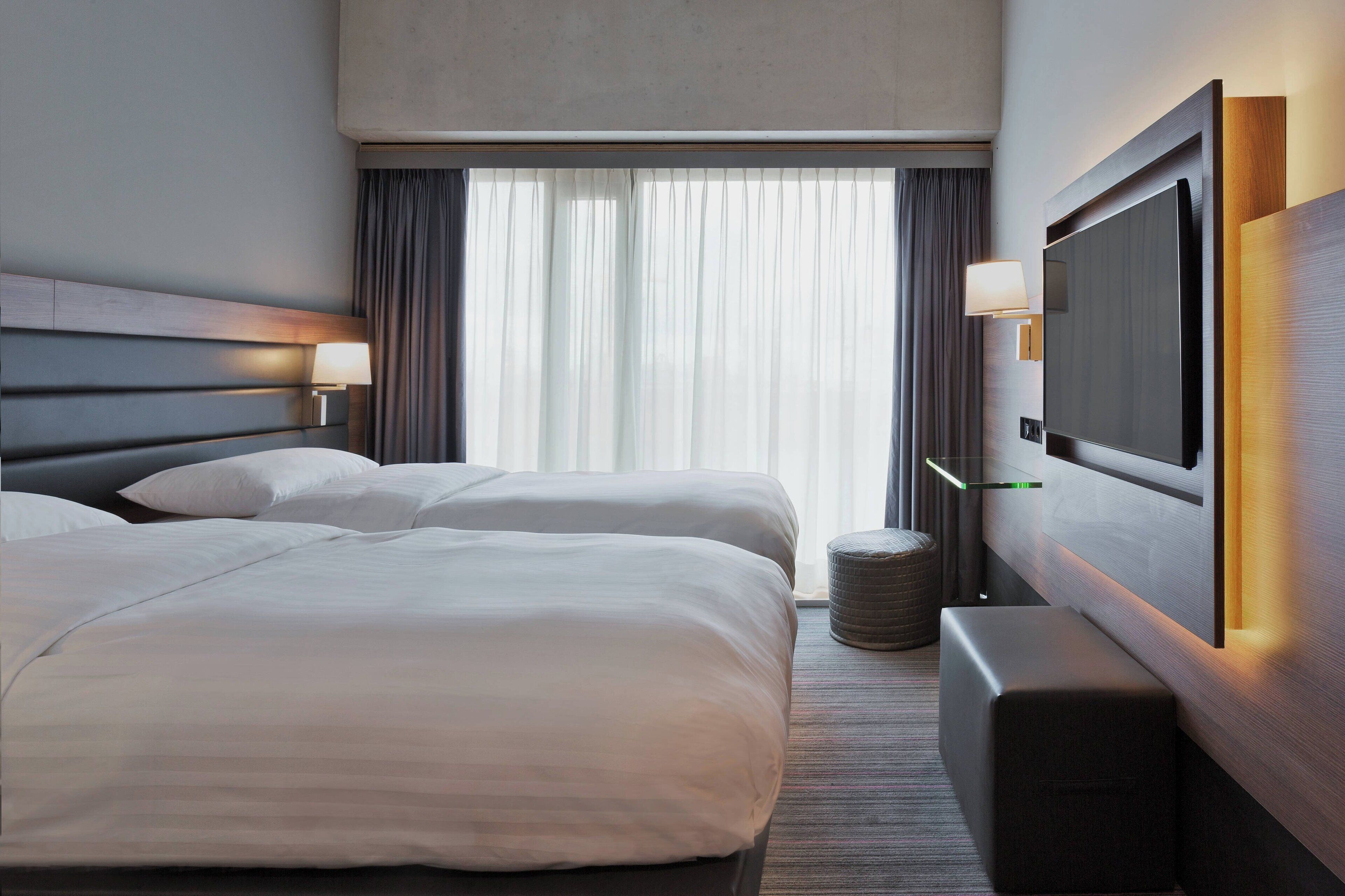 Habitación de hotel con dos camas, un televisor montado en la pared y un ventanal con cortinas.