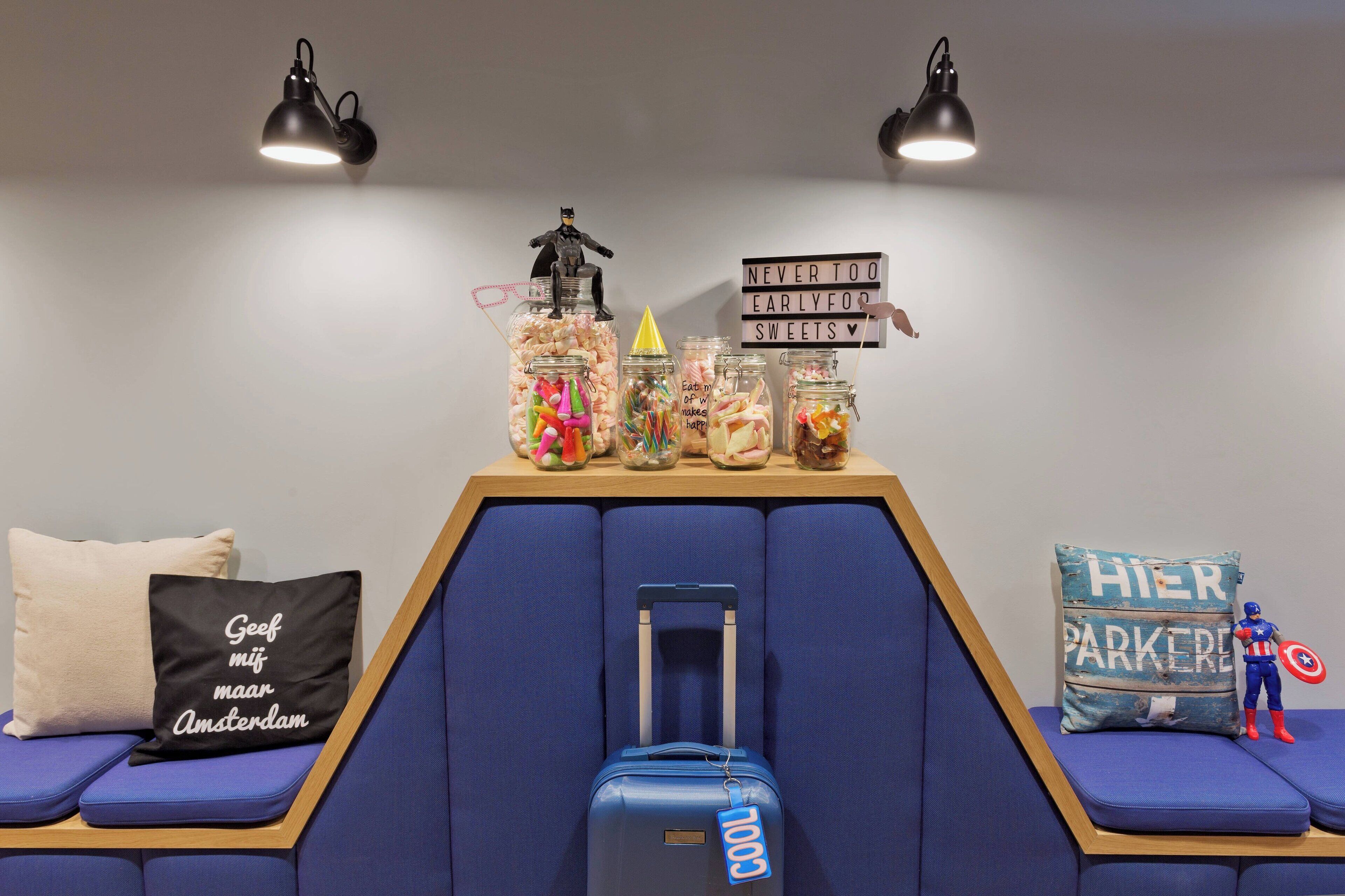 A hotel room with a blue sofa, a small table with snacks and a Batman figurine, a sign saying "NEVER TOO EARLY FOODS SWEETS", and a blue suitcase with a "COOL" tag.