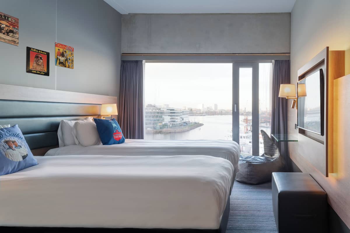 Quarto de hotel com duas camas, uma janela ampla com vista para a paisagem urbana e para a água, e um espaço de estar com uma luminária e uma televisão.