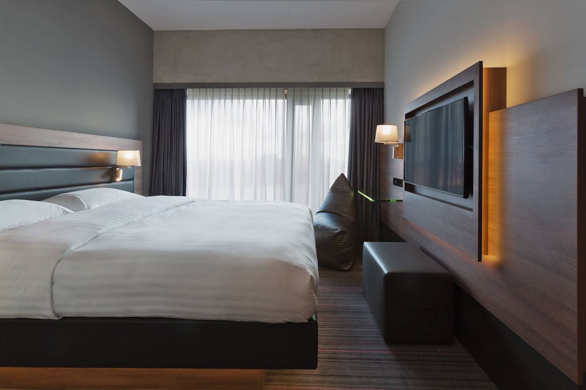 Quarto de hotel com uma cama grande, televisão de tela plana, mesa de cabeceira, uma cadeira e uma janela com cortinas.