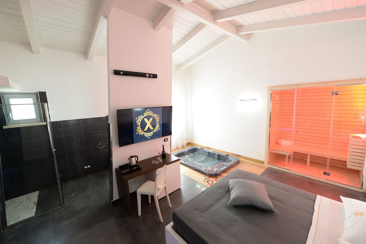 A modern hotel room with a bed, a desk, a TV, a sauna, and a shower area.