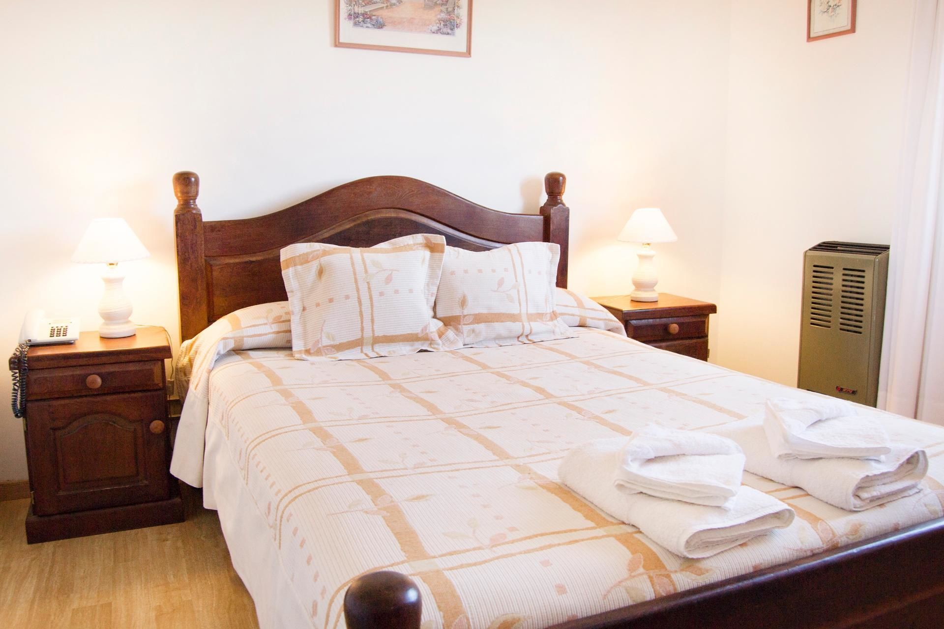 A neatly made bed with a patterned bedspread, two bedside lamps, a bedside telephone, and a heater on the right.