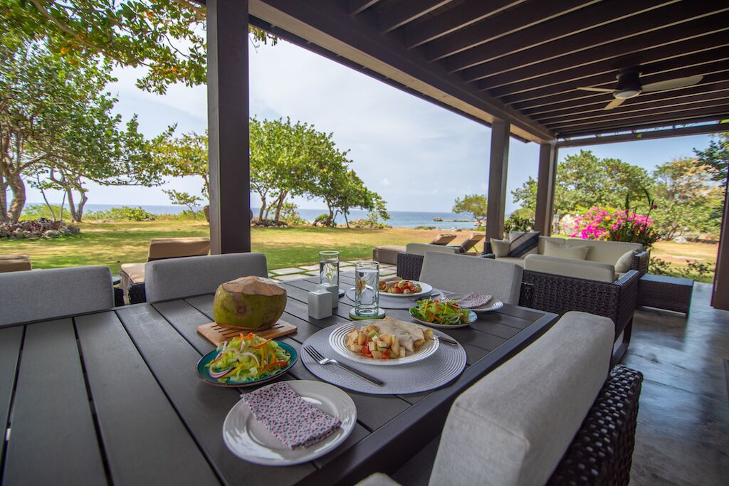 Un área de comedor al aire libre, techada y con vista al océano, que incluye una mesa dispuesta con platos de comida, un coco y un vaso de agua.