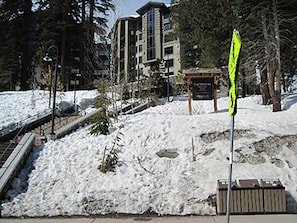 Un espacio exterior cubierto de nieve con un banco de madera, un cartel señalizador y un edificio al fondo.