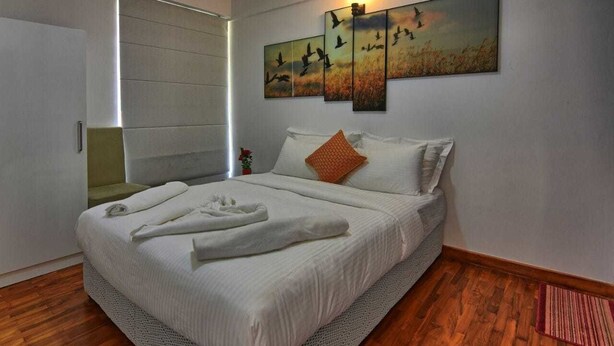 Un lit bien fait, avec du linge de lit blanc et un seul coussin orange, une œuvre d’art accrochée au mur représentant des oiseaux et un plancher en bois.