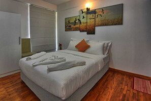 A neatly made bed with white linens and a single orange pillow, a wall-mounted artwork depicting birds, and a wooden floor.