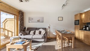Un salon moderne avec un canapé, une table basse et un coin repas. Il y a une grande fenêtre avec des rideaux, un tableau au mur et des meubles en bois dans la cuisine.