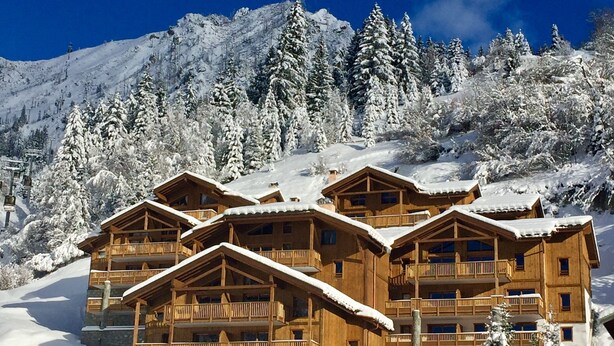 Un chalet en bois à plusieurs étages, doté de balcons, entouré d’arbres et de montagnes enneigées.