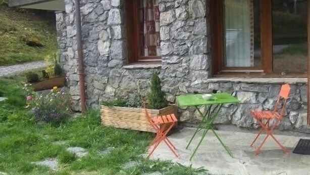 Een stenen gebouw met een kleine tuin, een houten terras en een groene tafel met twee oranje stoelen.