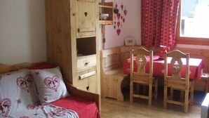 Een gezellige kamer met een houten garderobe, een bed met kussens met hartjesprint, een eettafel met stoelen en een raam met rode gordijnen.