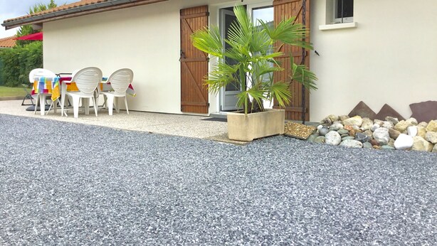 A building with a tiled roof, a gravel driveway, a wooden door, and a small window. There are white plastic chairs and a colorful tablecloth outside. - Maisonette 2 persons   (Biscarrosse)