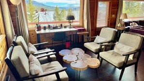 Interior de una cabaña con escritorio, sillas, una mesa redonda de madera y vistas a un lago y montañas.