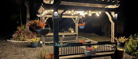 A wooden pergola with hanging plants and string lights, surrounded by potted plants and a bench.