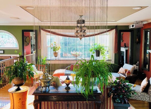 A cozy living room with a large window, a chandelier, and various plants.