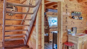 Interior de una cabaña de madera con una escalera de madera, una zona de cocina con estantes y utensilios, y un taburete rojo.