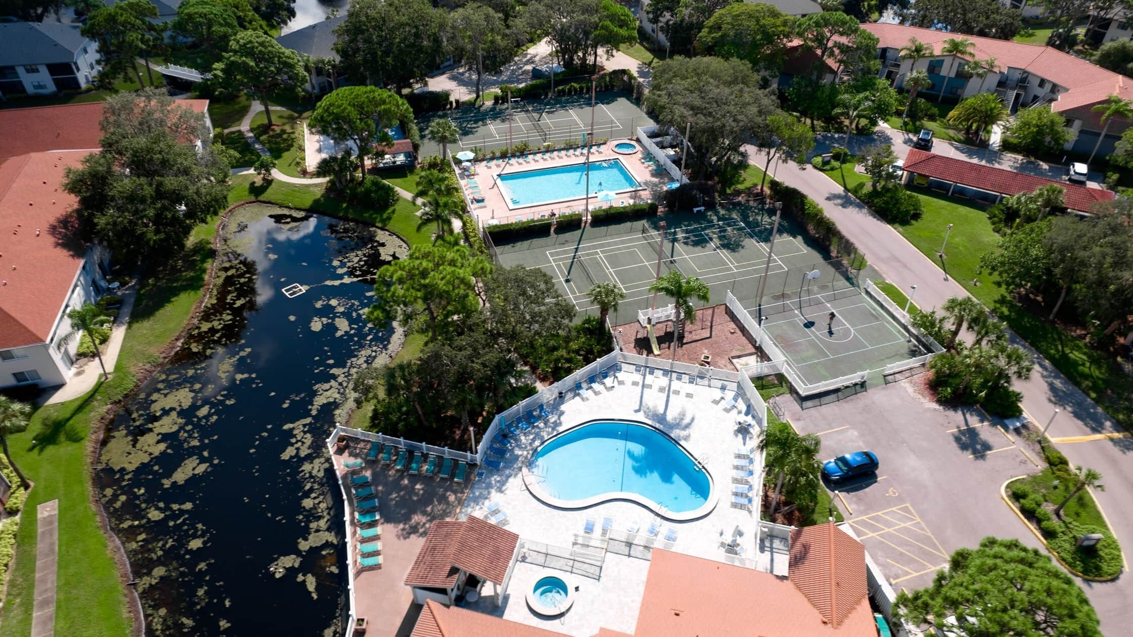 Vue aérienne d’un quartier résidentiel avec une piscine, des courts de tennis et un petit quai au bord d’un plan d’eau.