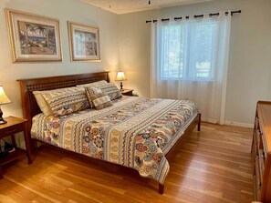 Un dormitorio con una cama de madera, dos mesitas de noche, un pequeño escritorio y una ventana con cortinas, además de dos cuadros en la pared.