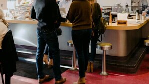 Interno di un bar con un bancone, una ventola a soffitto e un cartello che dice 'caffè buono, persone gentili, si prega di ordinare qui'.