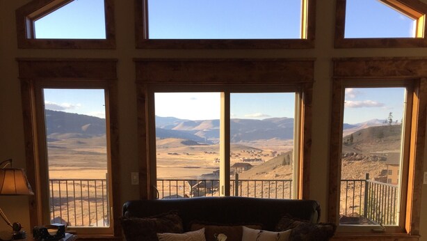 Una habitación con un gran ventanal con vistas a un paisaje montañoso. Hay un sofá de cuero marrón oscuro con cojines blancos y un jarrón con flores blancas sobre una mesa de centro de madera.