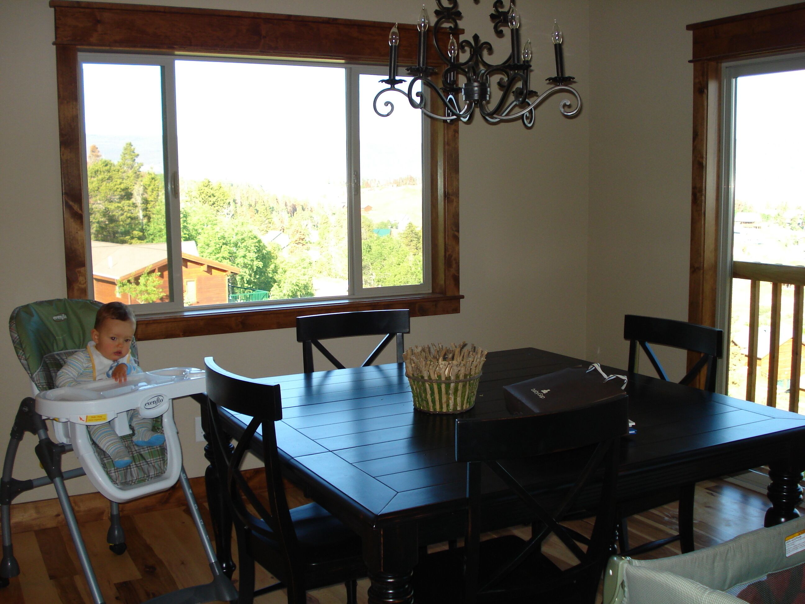 A dining area with a high table and chairs, a baby in a high chair, and a view of greenery outside.