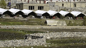 Une rangée de tentes surmontées de toiles blanches, disposées en ligne sur un terrain herbeux, avec des murs de pierre en arrière-plan.