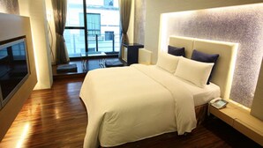 Quarto de hotel com uma cama grande, televisão, secretária com telefone e vista para a cidade pela janela.
