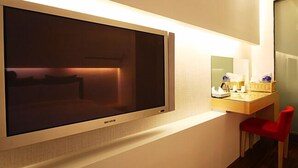 Quarto de hotel com uma televisão de tela plana grande fixada na parede, uma pequena escrivaninha com abajur e uma cadeira vermelha.
