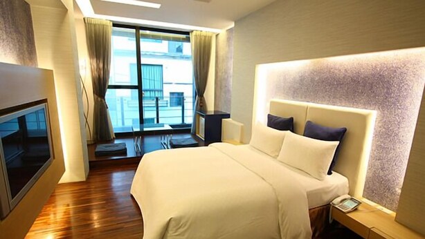 Quarto de hotel com uma cama grande, televisão, mesa de cabeceira com telefone e vista exterior através de uma janela ampla.