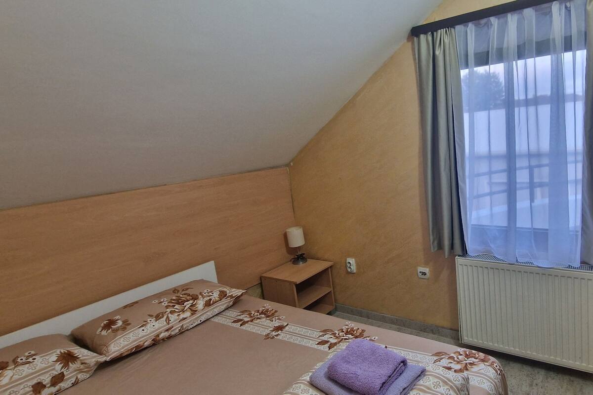 A bedroom with a bed, a nightstand, a window with curtains, and a radiator.