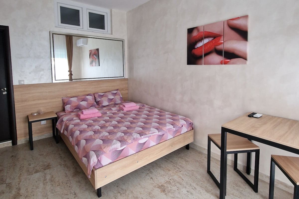 A bedroom with a bed, a desk, and a chair. There is a window with curtains, a wall-mounted air conditioner, and a painting of lips.
