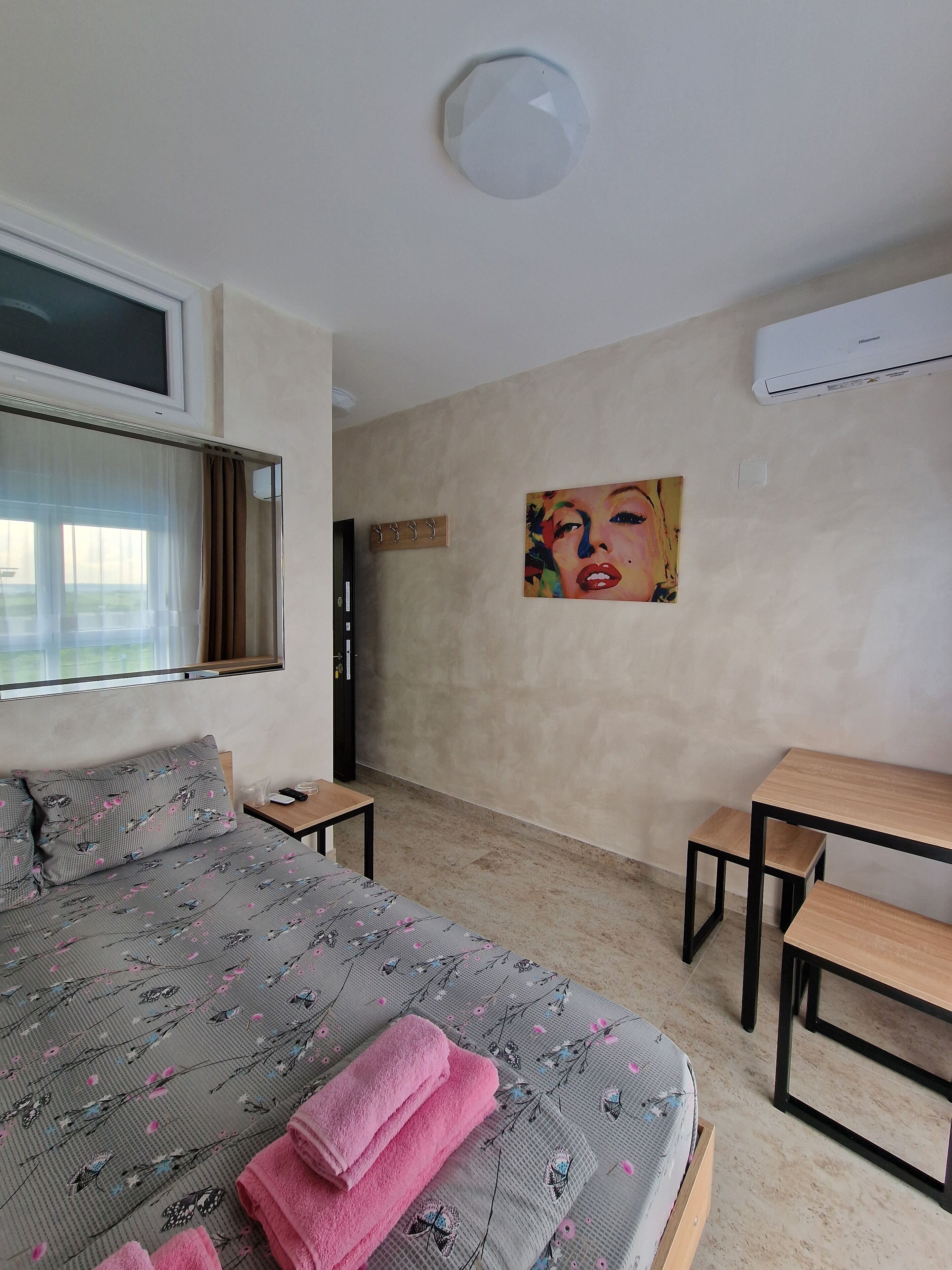 Une chambre à coucher comprenant un lit, un bureau, une chaise, une fenêtre avec des rideaux, un tableau au mur et un appareil de climatisation.