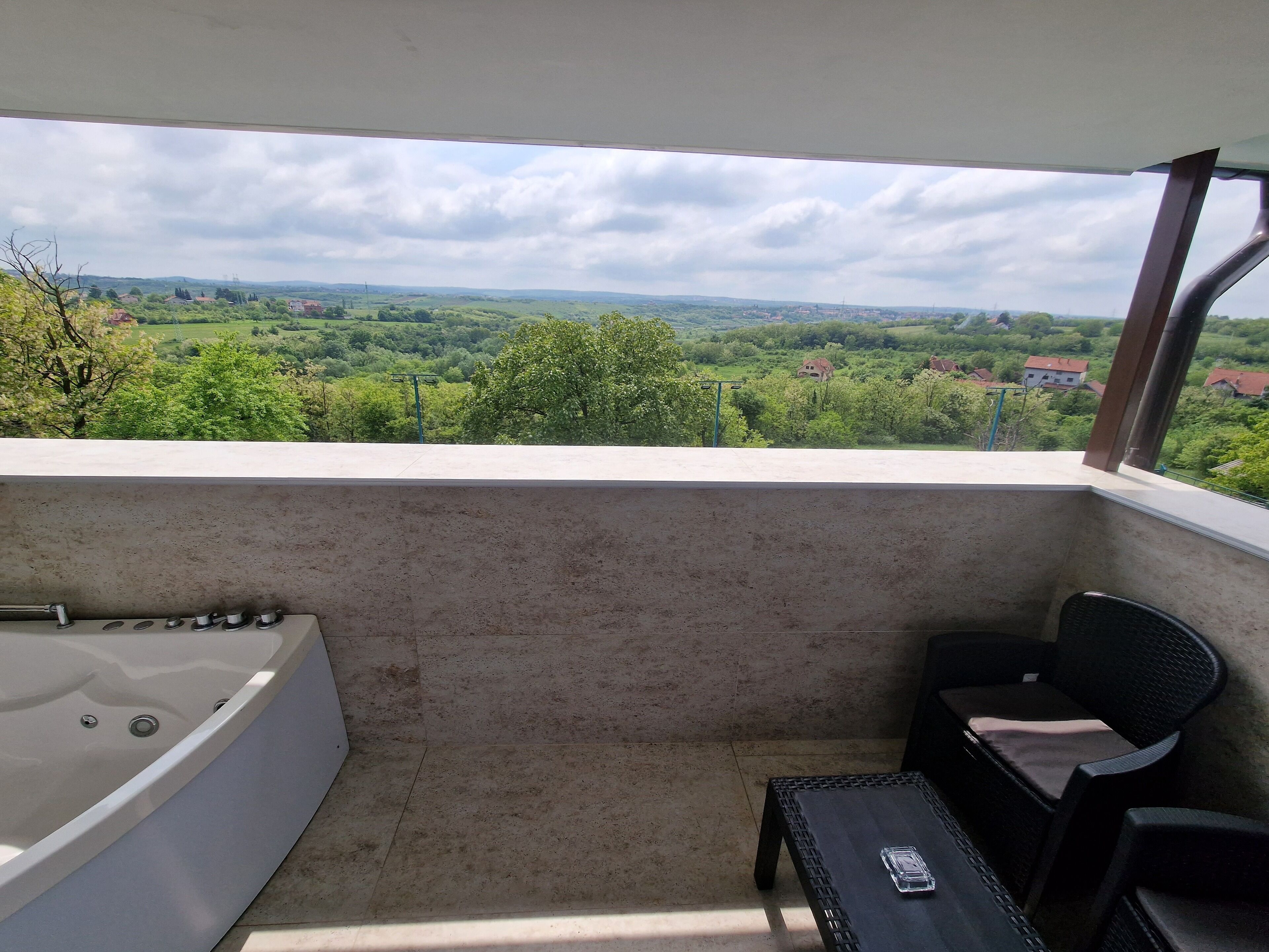 A balcony with a view of a green landscape, a bathtub, and two chairs.