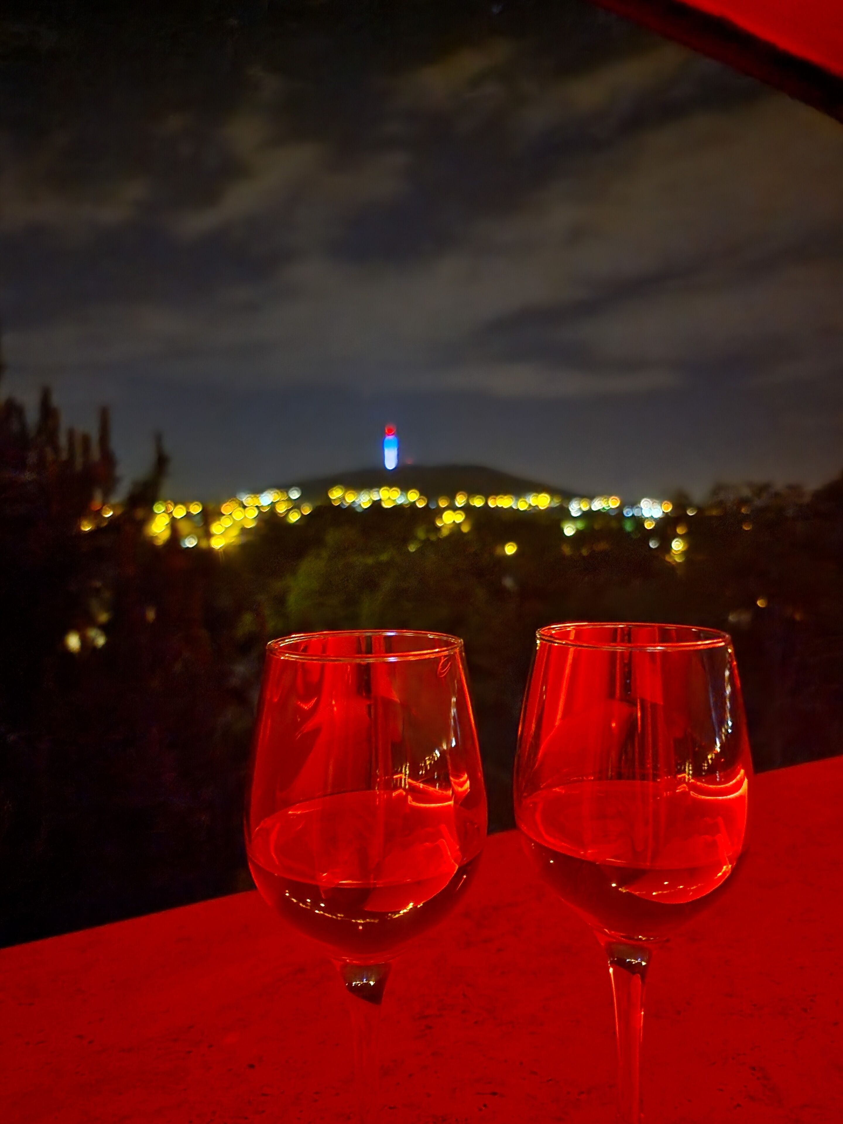 Twee lege wijnglazen op een rode tafel, met uitzicht op een stadsgezicht 's nachts.