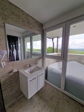 A bathroom with a large mirror, a sink, and a bathtub. There is a view of greenery outside through the windows.
