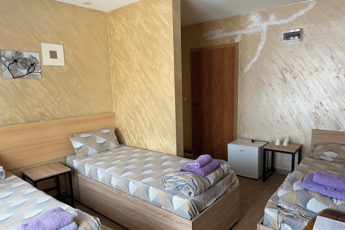 A hotel room with a wooden bed, a small desk, and a nightstand. There is a door leading to another room and a wall-mounted light fixture.