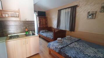 A compact room with a kitchenette, two single beds, a wooden cabinet, and a small shelf with glasses.