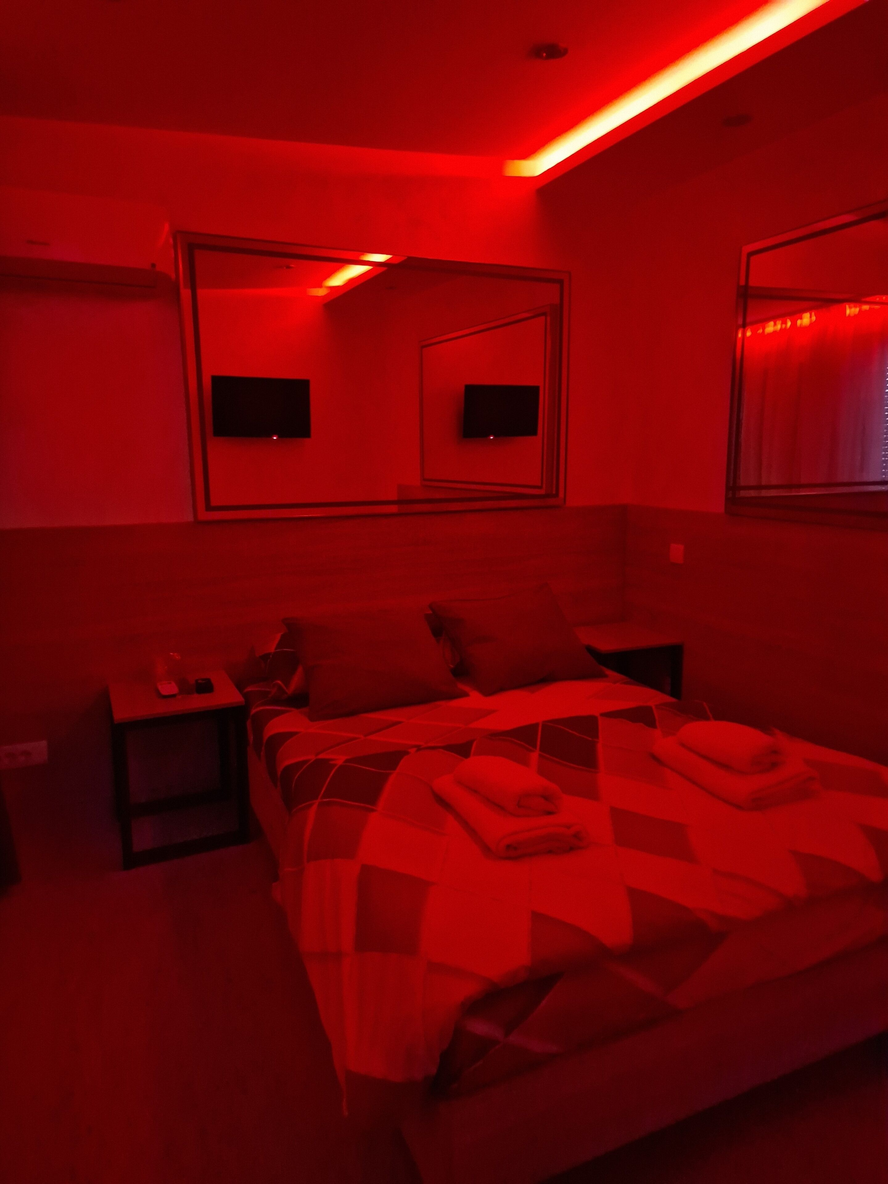 A dimly lit room with a red hue, featuring a bed with a checkered blanket, a nightstand, and two wall-mounted televisions.