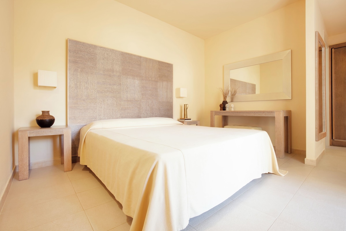 Ein Hotelzimmer mit einem großen Bett, einem Nachttisch, einem Spiegel und einer kleinen Vase.