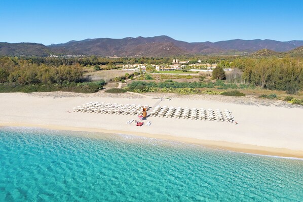 An aerial view of a beach with clear turquoise waters, a sandy shore, and a backdrop of mountains. - Spiagge San Pietro Resort (Castiadas)