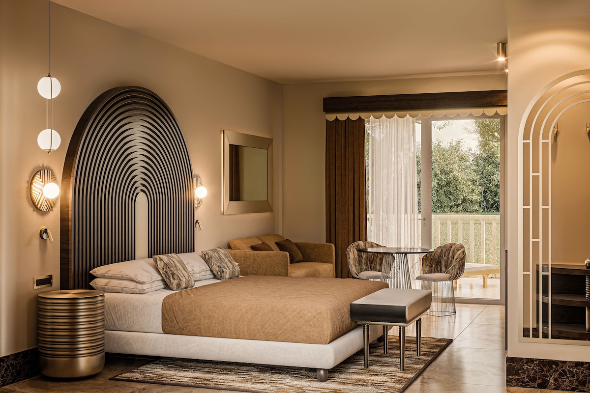 A modern bedroom with a large bed, a round side table, and a chair. There is a balcony with a view of greenery.