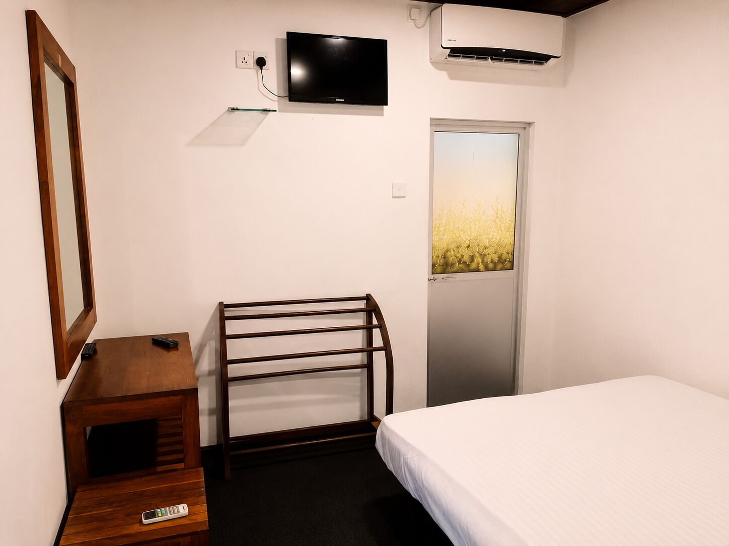 A hotel room with a bed, a TV mounted on the wall, an air conditioning unit, a door with a frosted glass panel, and a wooden nightstand with a remote control.
