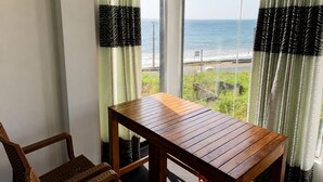 A room with a wooden table and a wicker chair, a large window with curtains, and a view of the ocean. - Villa Yuvin (Dondra)