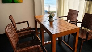 A room with a wooden table and wicker chairs, a painting of trees, and a view of the ocean. - Villa Yuvin (Dondra)