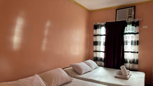 A room with two beds, each with a pillow and a rolled towel. There is a window with striped curtains and a radiator above it. - A's Azotea de Bohol (Tagbilaran)