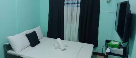 Uma cama de solteiro com roupa de cama branca, cortinas preto e branco, uma televisão na parede e uma pequena prateleira com objetos.