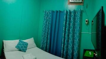 A room with teal walls, a bed with white bedding and a blue patterned pillow, a window with blue curtains, and a small air conditioning unit above the window.