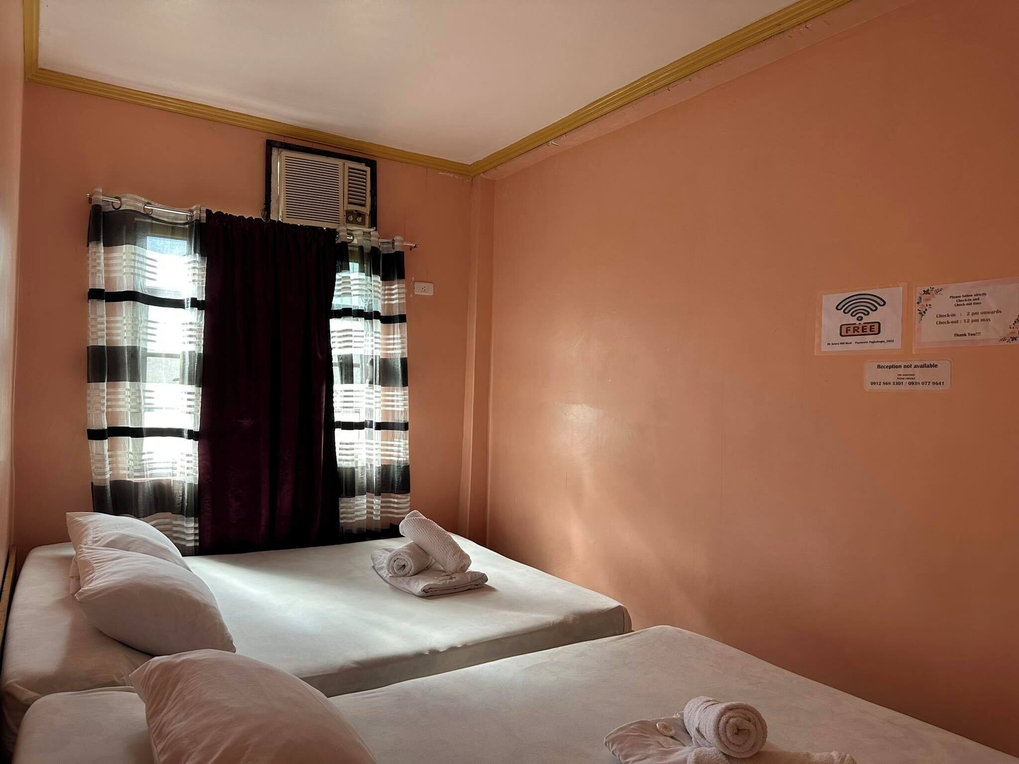 A hotel room with two beds, a window with curtains, a wall-mounted sign, and a reception sign indicating check-in times.